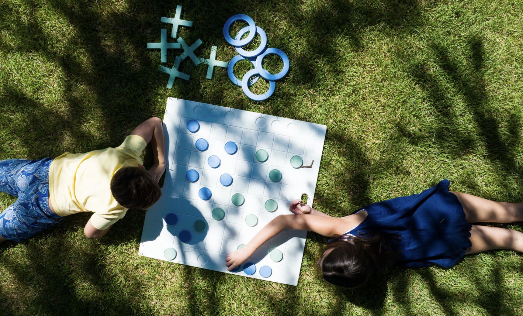 Two children in a vibrant scene play an immersive game of Checkers & Tic Tac Toe by Ledge Lounger on the grass. One wears a yellow shirt, the other a blue dress. The board displays scattered pieces, extra circles, and crosses, with tree-filtered sunlight enhancing the playful atmosphere.