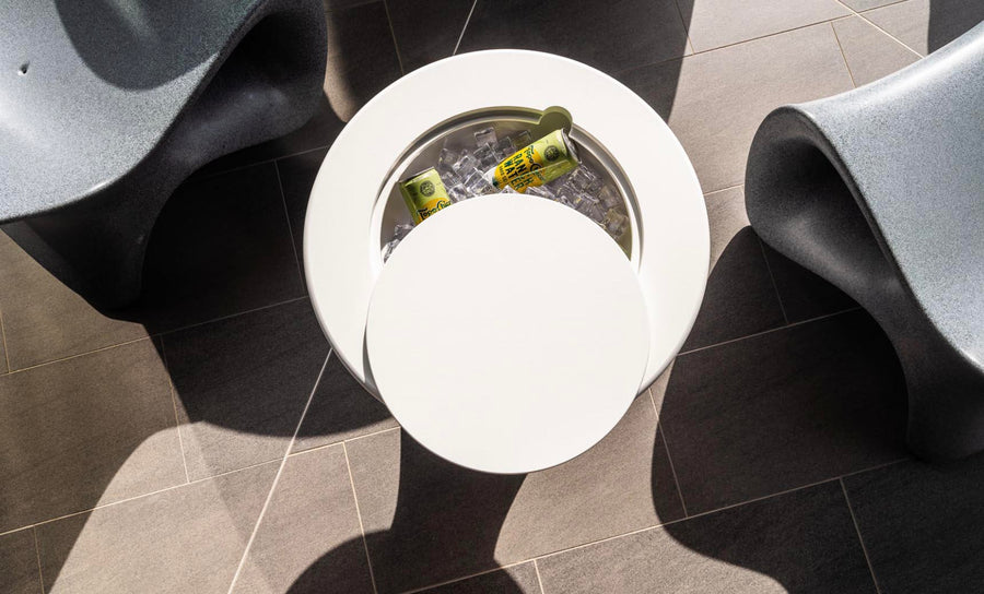 An Autograph Tall Ice Bin Side Table by Ledge Lounger, white and round, sits on a gray tiled floor with its lid open to reveal chilled canned drinks. Two modern curved gray chairs flank this versatile piece as sunlight casts playful shadows across the scene.