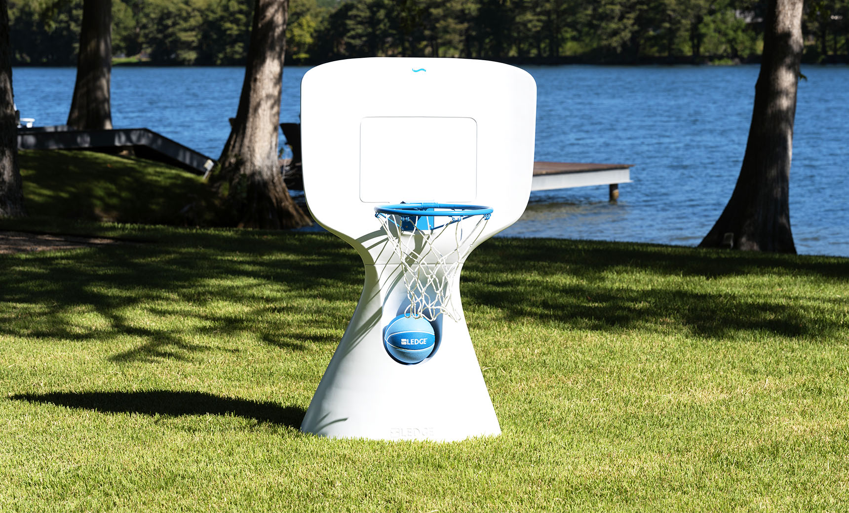 A modern Hoopstr basketball hoop by Ledge Lounger, featuring a blue rim and net, stands on a grassy lawn by the lake. Ideal for a backyard game, trees and a dock accentuate the scene under the clear blue sky.