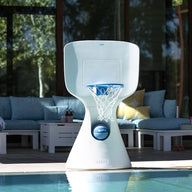 A modern white Ledge Lounger Hoopstr basketball hoop graces the poolside, set against a luxurious outdoor seating area with blue cushions. Sunlight casts shadows, enhancing the peaceful atmosphere, ideal for an impromptu backyard game.