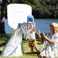 A child in yellow shorts happily shoots a blue basketball into the Ledge Lounger Hoopstr as a woman smiles and claps from the grass. Their backyard game unfolds lakeside, surrounded by trees under a clear sky.