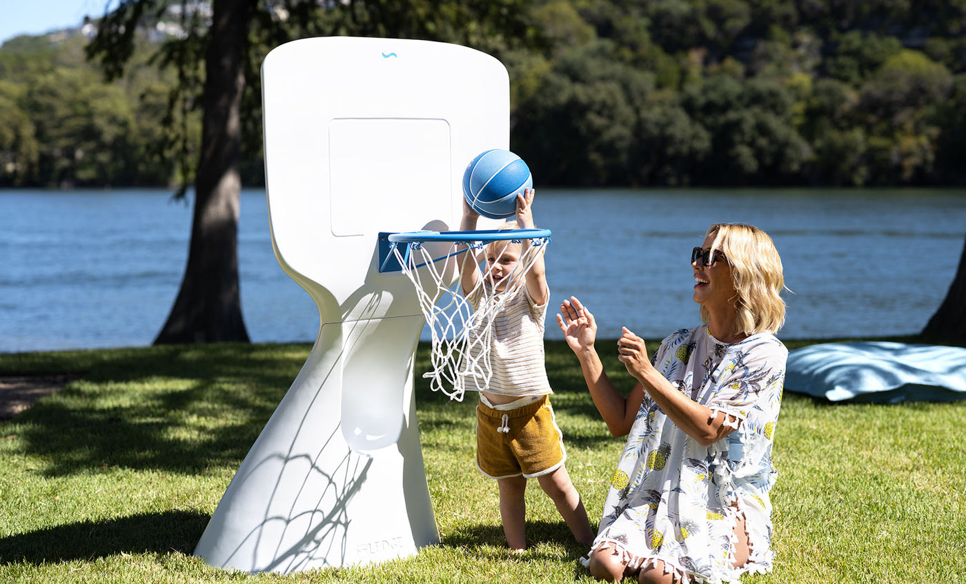 A child in yellow shorts happily shoots a blue basketball into the Ledge Lounger Hoopstr as a woman smiles and claps from the grass. Their backyard game unfolds lakeside, surrounded by trees under a clear sky.