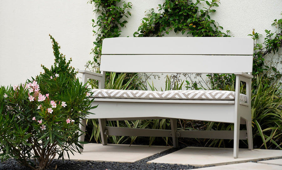A gray outdoor bench with a Mainstay Bench Cushion by Ledge Lounger, showcasing vibrant fabric colors, sits on a patio. Its surrounded by lush green plants, including a pink-flowered bush on the left. Ivy climbs the cream-colored wall behind it.