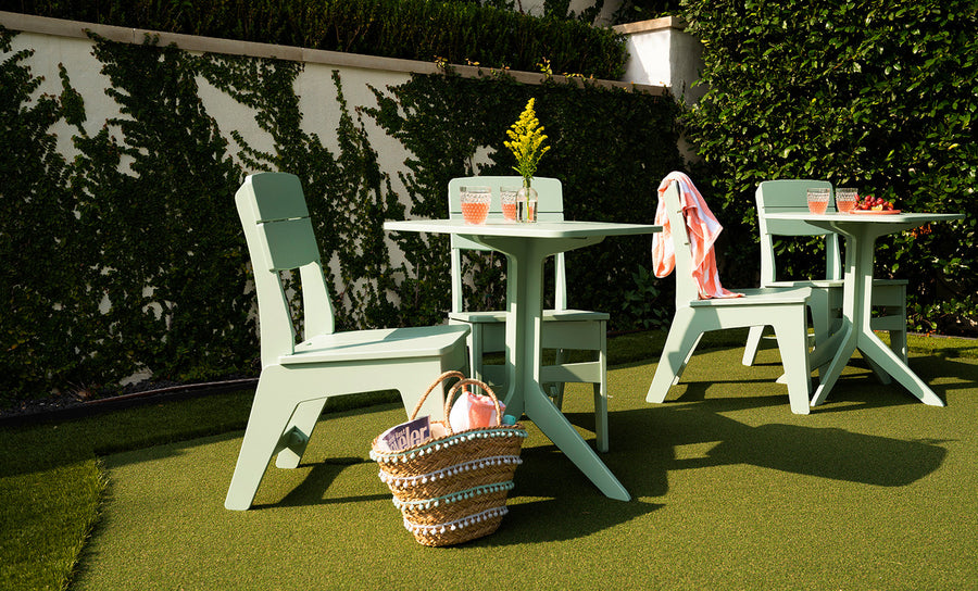 Outdoor patio with two mint green tables and chairs on a green lawn. Each table has drinks and flowers. A woven basket with a magazine is in the foreground, and a towel is draped over a chair. A hedge and wall are in the background.