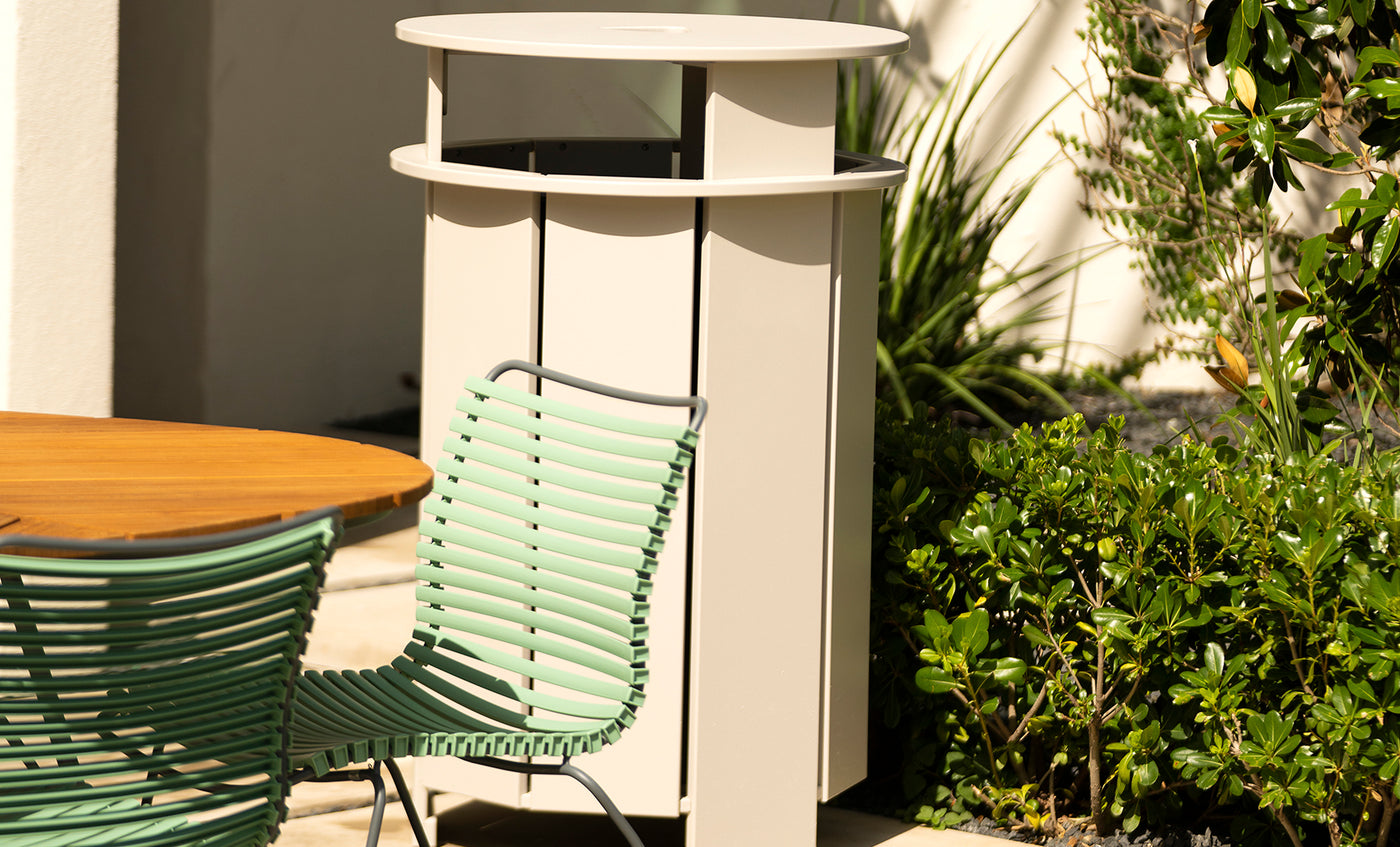 A modern patio scene includes a light green slatted chair beside a round wooden table. A sleek, light outdoor heater is surrounded by lush plants in the background, with sunlight casting gentle shadows nearby.