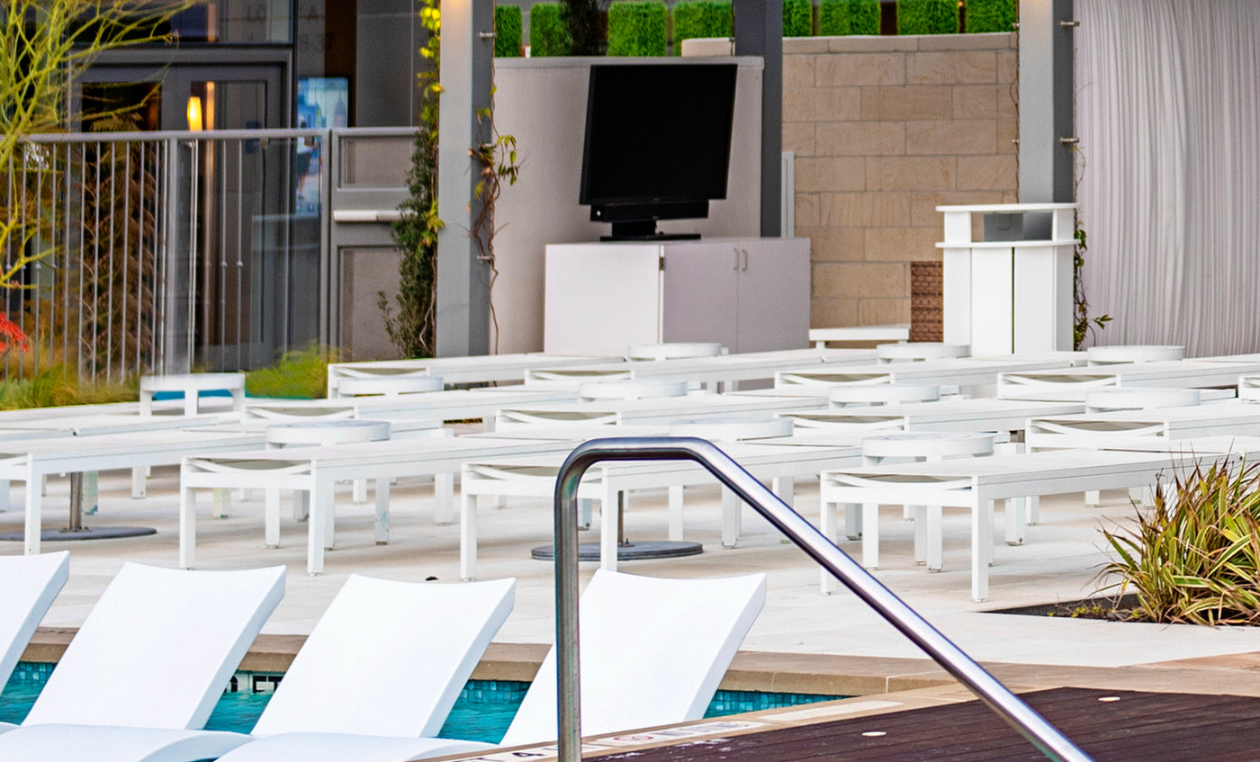 A contemporary outdoor poolside features white Ledge Lounger chairs facing a TV on a stand, surrounded by potted plants, a stainless steel railing, and a dry bar under a roof with wooden decking in the foreground.