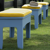 Three Mainstay Dining Benches, 25 by Ledge Lounger, with blue frames and yellow striped cushions are arranged on green grass near a black wooden fence. A tan hat is placed on the bench in front.