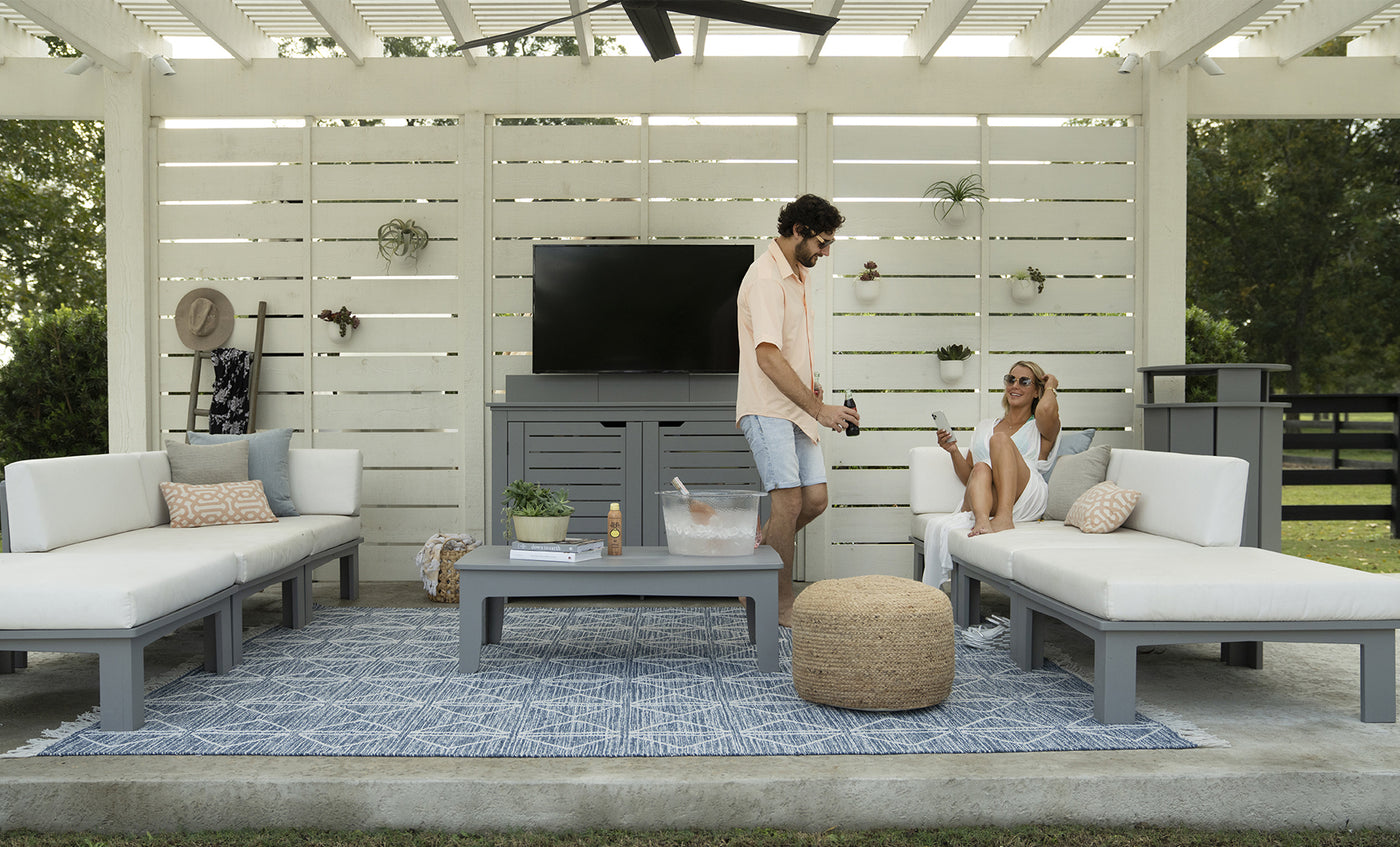A man and woman relax on outdoor white sofas with a patterned rug; he serves drinks from an ice chest on the table. The area features a white slatted backdrop, wall-mounted TV, and decorative straw hat. Brand products visible: Ledge Lounger (no Mainstay Trash Bin in sight).