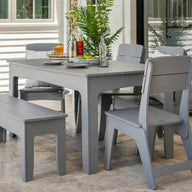 The setup includes a gray patio table with matching chairs and the Mainstay Dining Bench 25 from Ledge Lounger. The table features tableware, a plant centerpiece, and a glass bottle. Large windows and potted plants complete the serene environment.