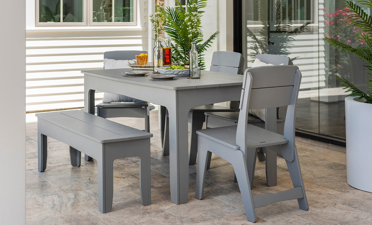 The setup includes a gray patio table with matching chairs and the Mainstay Dining Bench 25 from Ledge Lounger. The table features tableware, a plant centerpiece, and a glass bottle. Large windows and potted plants complete the serene environment.