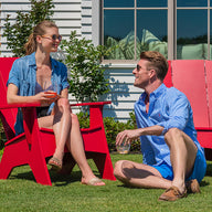A woman sits on a Mainstay Ultimate Adirondack from Ledge Lounger, drink in hand, while a man relaxes nearby. Both don sunglasses and casual summer wear, enjoying the sunny garden bliss with chic outdoor lounge chairs in front of their houses large windows.