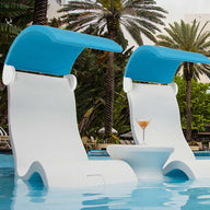 Two white Ledge Lounger Signature Chair Shades with blue canopies sit on the in-pool tanning ledge, partially submerged. Between them, a small round table holds a cocktail. Tall palm trees and a white building create a serene backdrop.