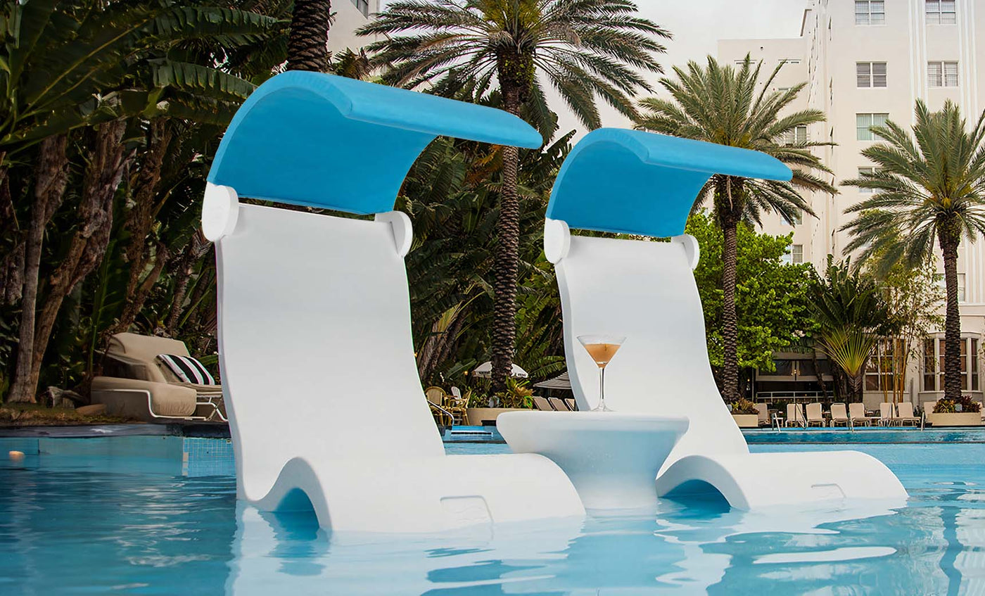 Two white Ledge Lounger Signature Chair Shades with blue canopies sit on the in-pool tanning ledge, partially submerged. Between them, a small round table holds a cocktail. Tall palm trees and a white building create a serene backdrop.