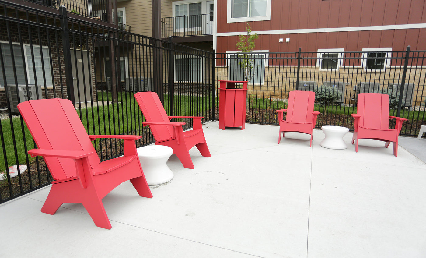 Four red Adirondack chairs form a semi-circle on a concrete patio, flanked by two white side tables. The space is bordered by a black metal fence, with apartment buildings and balconies in view.
