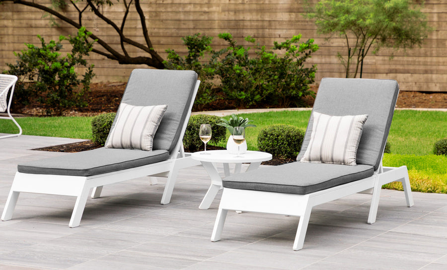 Two outdoor lounge chairs with gray cushions and Ledge Loungers Throw Pillow 18 Square - Final Sale are on a patio. A small white table between them holds a wine glass and plant, with green shrubs and a wooden fence in the background.