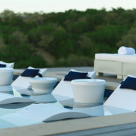The modern poolside features the Ledge Lounger Signature Chaise and Affinity Side Table In-Pool Set, with white loungers partially in water, round side tables, a white outdoor couch with navy cushions and towels, all framed by lush trees.