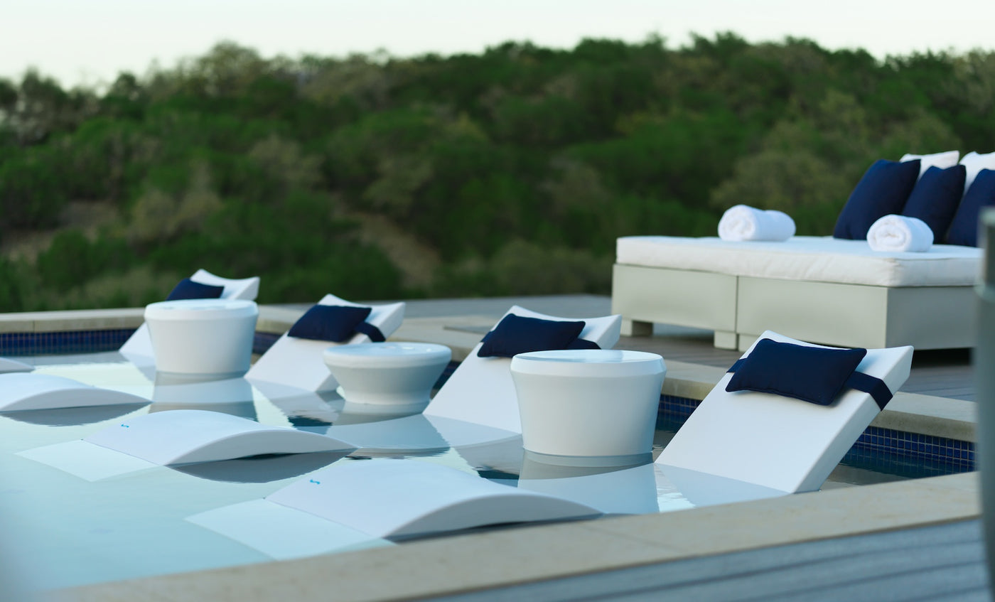 The modern poolside features the Ledge Lounger Signature Chaise and Affinity Side Table In-Pool Set, with white loungers partially in water, round side tables, a white outdoor couch with navy cushions and towels, all framed by lush trees.