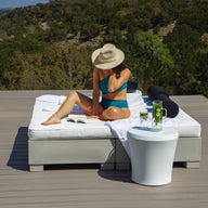 A woman in a blue bikini and sunhat relaxes on a lounge bed, reading by the Affinity Side Table from Ledge Lounger. Drinks rest atop the table as she enjoys nature and sunshine on a wooden deck surrounded by trees.
