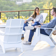 A woman and a man relax on Mainstay Ultimate Adirondack chairs by Ledge Lounger on the patio, sipping wine. A fire pit flickers between them, with lush greenery and string lights in the background, creating a serene atmosphere.