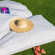 A straw hat and a magazine rest on Ledge Loungers Laze Pillow with a subtle pattern, crafting an idyllic scene on the lush green grass. Nearby, orange sandals featuring red straps complete the outdoor comfort vibe.