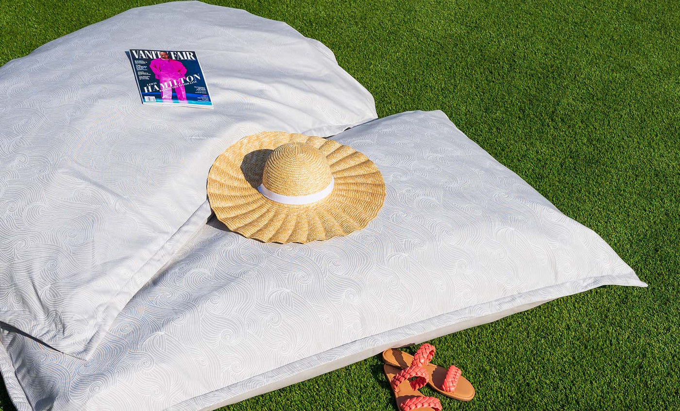 A straw hat and a magazine rest on Ledge Loungers Laze Pillow with a subtle pattern, crafting an idyllic scene on the lush green grass. Nearby, orange sandals featuring red straps complete the outdoor comfort vibe.