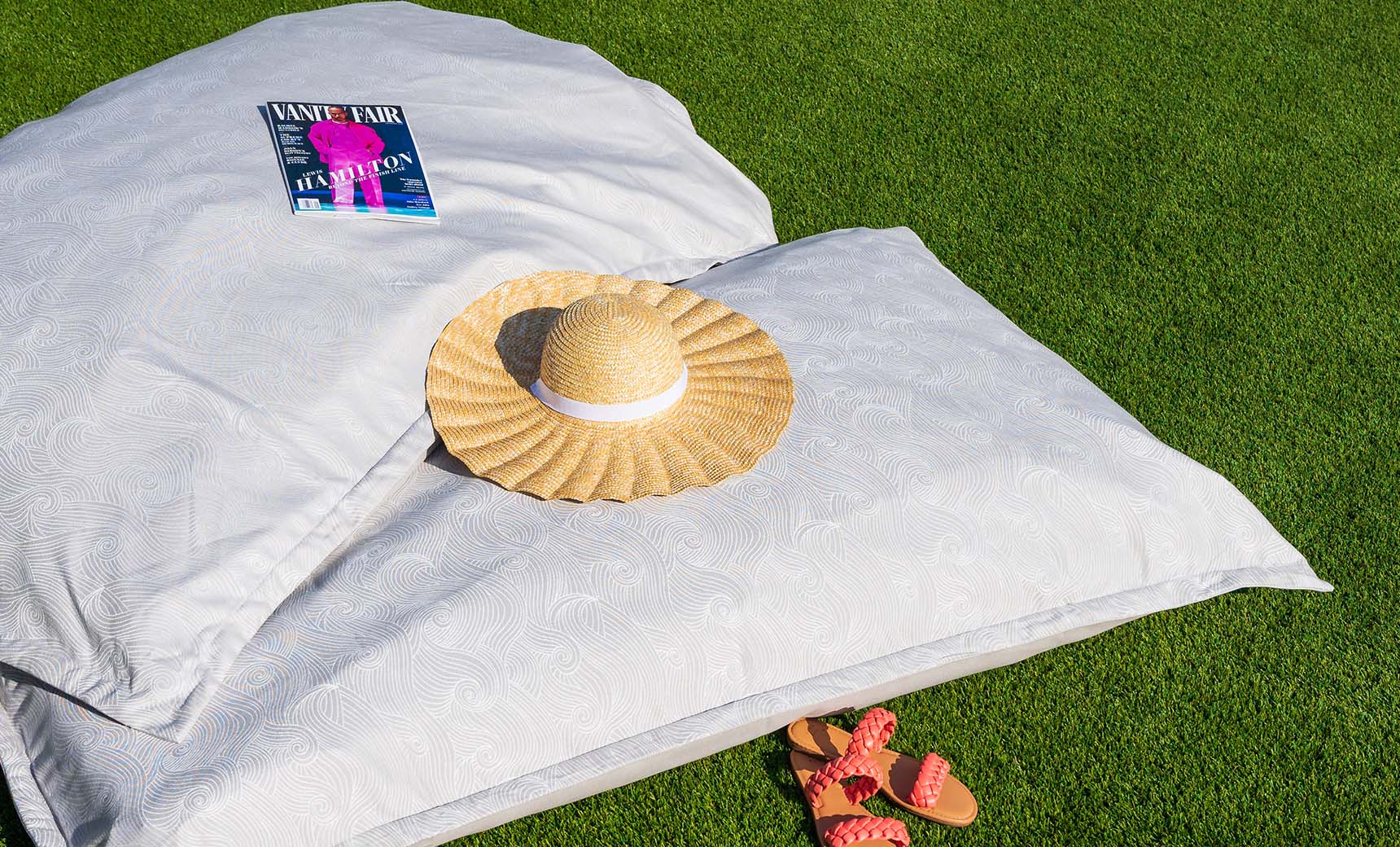 A straw hat and a magazine rest on Ledge Loungers Laze Pillow with a subtle pattern, crafting an idyllic scene on the lush green grass. Nearby, orange sandals featuring red straps complete the outdoor comfort vibe.
