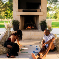 Two people relax on Ledge Loungers Laze Pillows, enjoying a board game on a glass table near the lit outdoor fireplace. The covered patio offers views of lush lawns and trees, enhancing the outdoor comfort of this cozy setting.