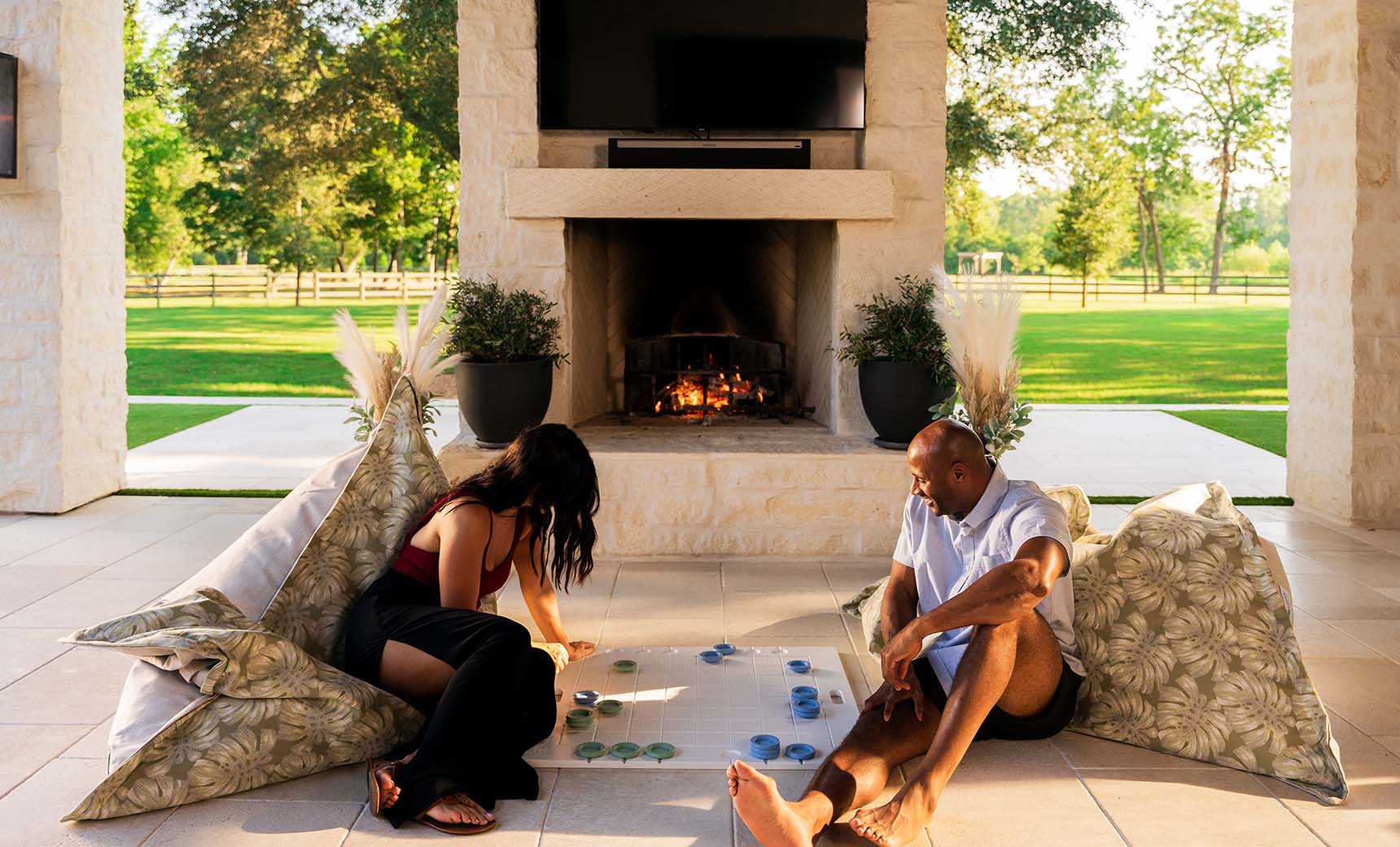 Two people relax on Ledge Loungers Laze Pillows, enjoying a board game on a glass table near the lit outdoor fireplace. The covered patio offers views of lush lawns and trees, enhancing the outdoor comfort of this cozy setting.