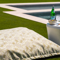 A large floral pillow is on a grassy lawn next to the Affinity Side Table by Ledge Lounger, which holds a sparkling water bottle, an ice-filled glass, and sliced fruit. In the background, a spacious swimming pool with clear water and an elegant tanning ledge is visible.