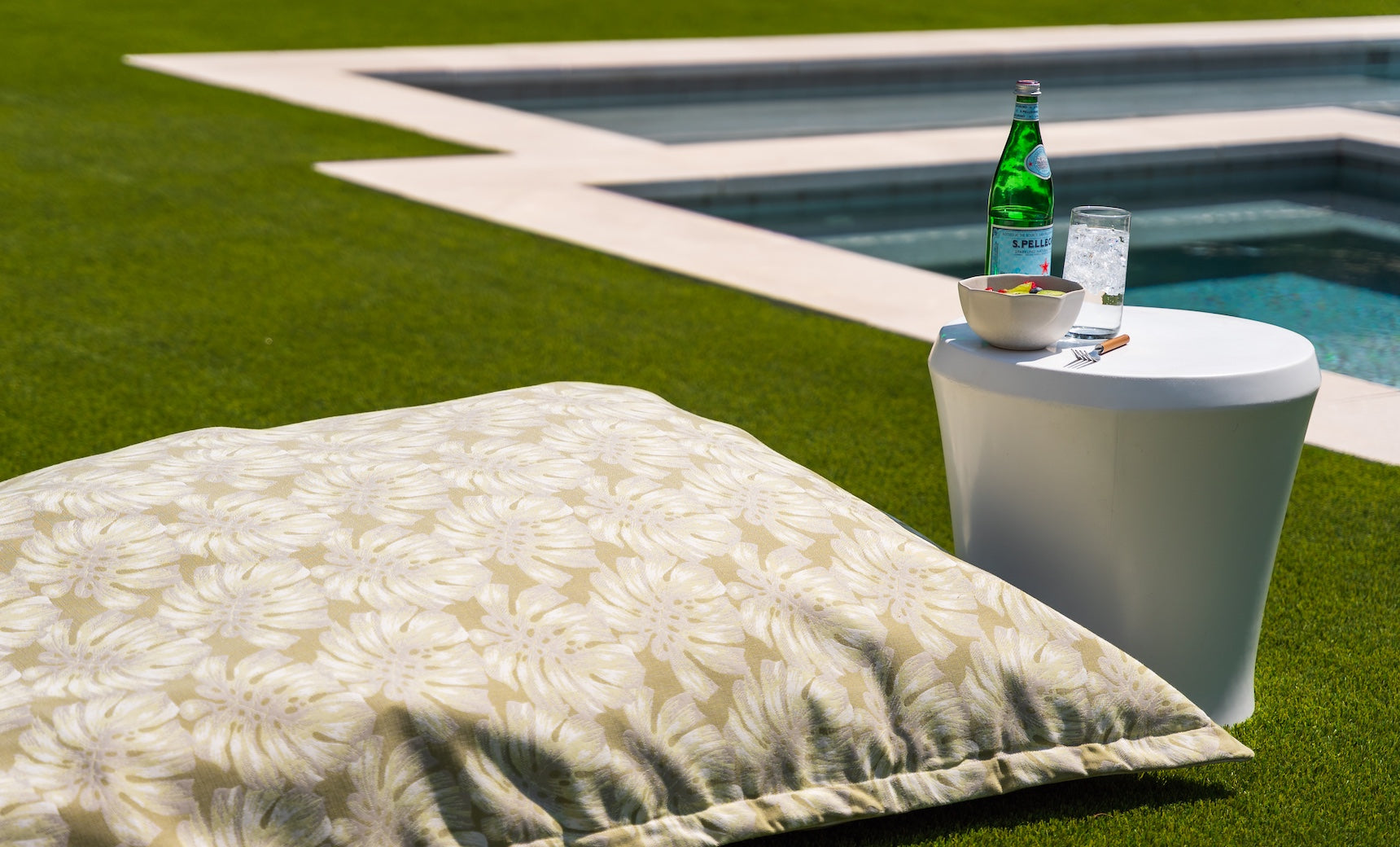 A large floral pillow is on a grassy lawn next to the Affinity Side Table by Ledge Lounger, which holds a sparkling water bottle, an ice-filled glass, and sliced fruit. In the background, a spacious swimming pool with clear water and an elegant tanning ledge is visible.