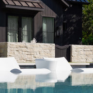 Three modern white Ledge Lounger Signature Chaise and Affinity Side Table In-Pool Set pieces are partially immersed on a pool ledge in front of a dark house with large windows, stone wall, and lush greenery—ideal for stylish poolside lounging.