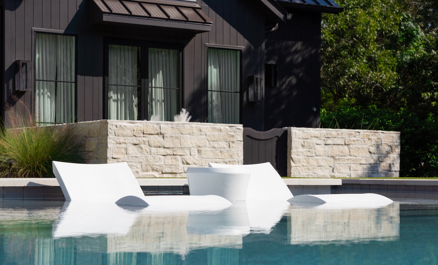 Three modern white Ledge Lounger Signature Chaise and Affinity Side Table In-Pool Set pieces are partially immersed on a pool ledge in front of a dark house with large windows, stone wall, and lush greenery—ideal for stylish poolside lounging.