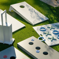 Ledge Lounger Cornhole and other outdoor yard games rest on green grass, featuring portable stackable boards with handles and playful tic-tac-toe sets. Sunlight casts lively shadows over the scene.