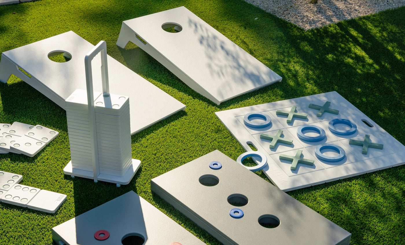Ledge Lounger Cornhole and other outdoor yard games rest on green grass, featuring portable stackable boards with handles and playful tic-tac-toe sets. Sunlight casts lively shadows over the scene.