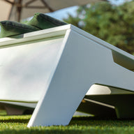 Close-up of a modern white outdoor lounge chair with two green cushions, set on artificial grass beside a Ledge Lounger Cornhole set. Blurred trees and parasols are in the background; ELLEFGE is engraved on the side.
