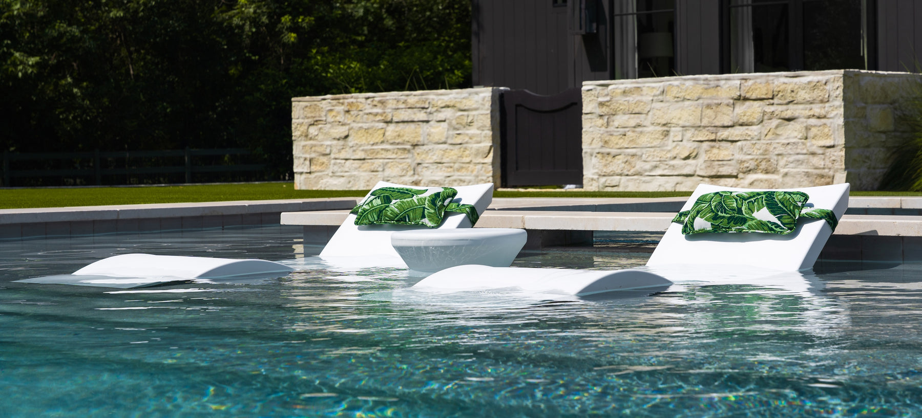 Two white lounge chairs partially submerged in a swimming pool with patterned green cushions. A small white table is between the chairs, and a stone wall with windows is visible in the background. Lush greenery surrounds the area.