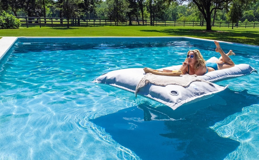 A woman in sunglasses relaxes on a floating pool mattress with a book, enjoying a sunny day in a clear blue swimming pool surrounded by green grass and trees.