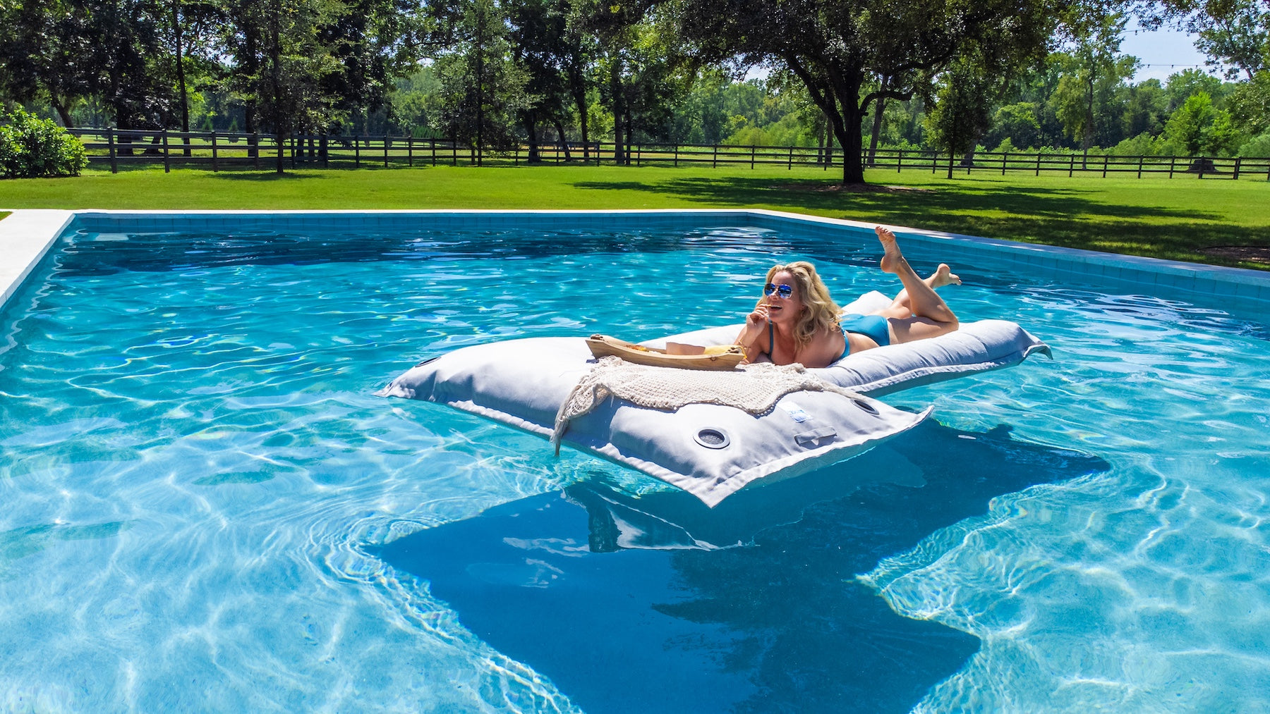 A woman relaxes on an inflatable pool mattress, wearing sunglasses and a swimsuit, in a clear blue outdoor pool surrounded by grass, trees, and a wooden fence on a sunny day.