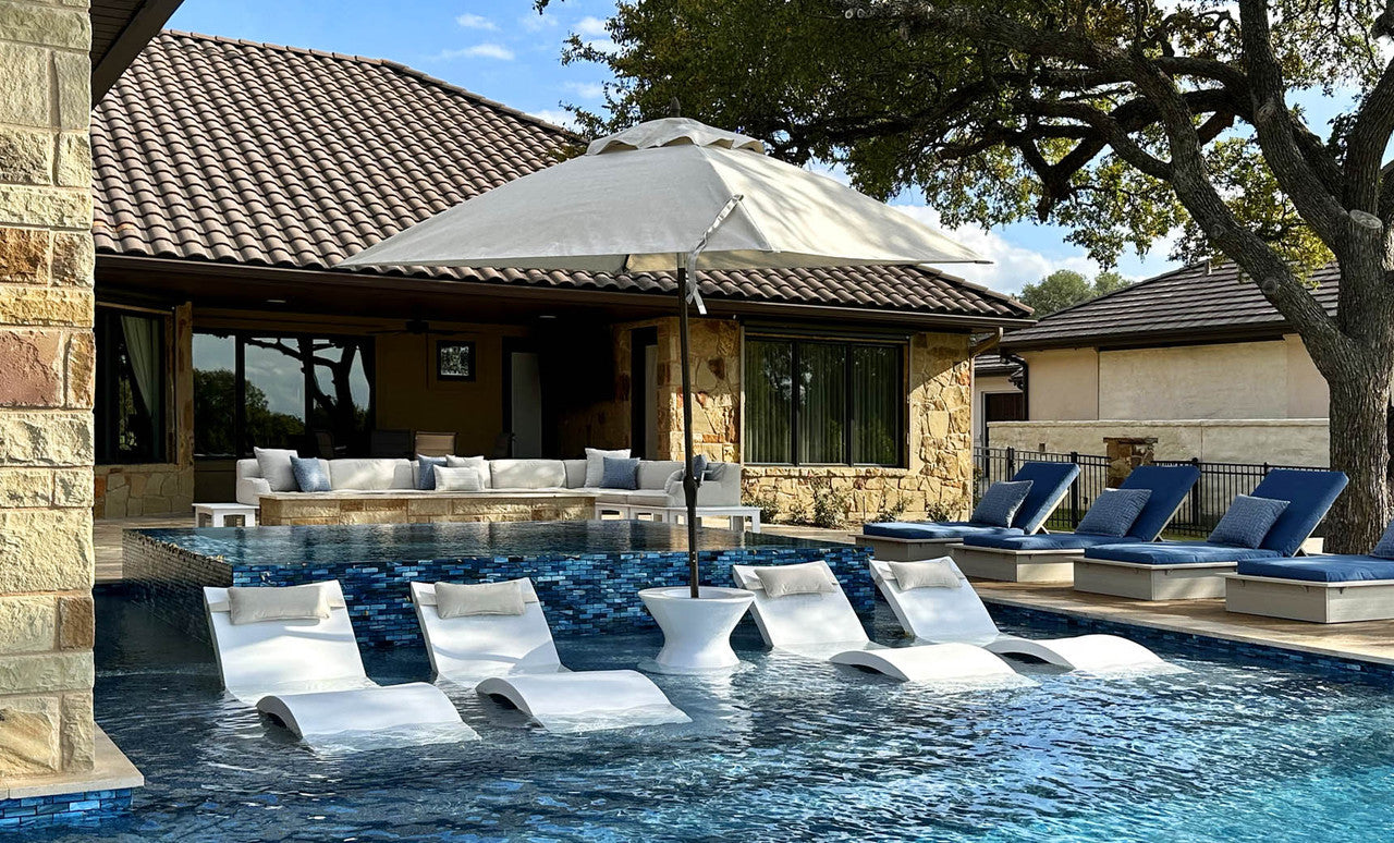 Luxurious outdoor pool with white lounge chairs partially submerged in water features the Essential Fiberglass Umbrella - 11 Octagon Pulley from Ledge Lounger for shade. The pool is bordered by stone buildings with tiled roofs, cushioned seating, and a backdrop of trees and a fence.
