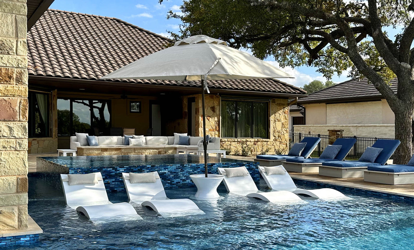A stone house and large swimming pool with submerged loungers and blue sunbeds create a luxurious backyard. The area is shaded by the Essential Fiberglass Umbrella - 10 Square Pulley from Ledge Lounger, and is surrounded by trees under clear skies for a serene atmosphere.