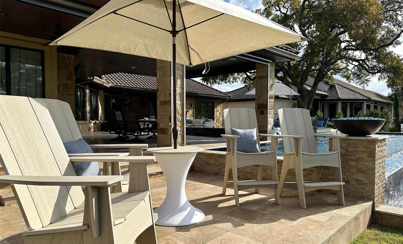 A patio scene features two wooden chairs and a small white table beneath the Essential Fiberglass Umbrella - 10 Square Pulley by Ledge Lounger, with marine-grade fabrics. A swimming pool and planters with greenery adorn the backdrop, framed by a stone house under a clear blue sky.