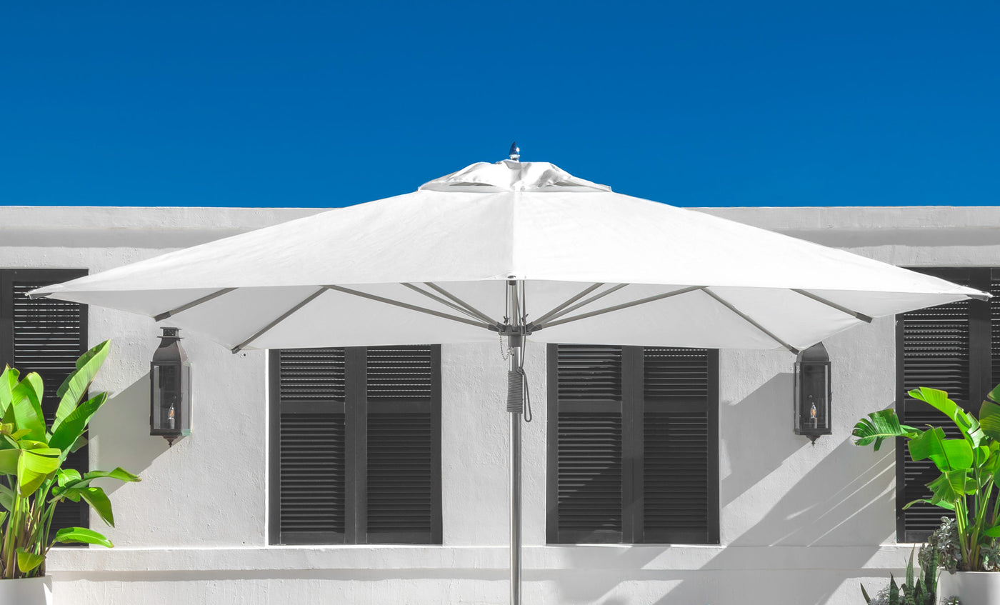 A 10 Square Pulley Essential Fiberglass Umbrella by Ledge Lounger stands open on a sunny day against a white building with dark shutters, framed by green plants and a clear blue sky, highlighting its durable fiberglass ribs and marine-grade fabrics.