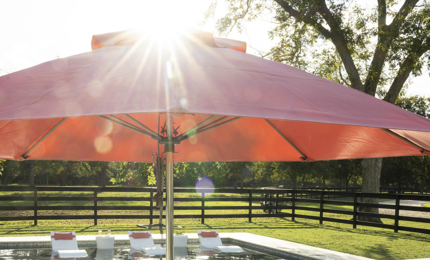 The Essential Fiberglass Umbrella - 10 Square Pulley by Ledge Lounger shades a poolside area with white lounge chairs. Sunlight filters through the trees and umbrella, casting a warm glow. Supported by sturdy bases, it complements the black wooden fence and lush greenery.
