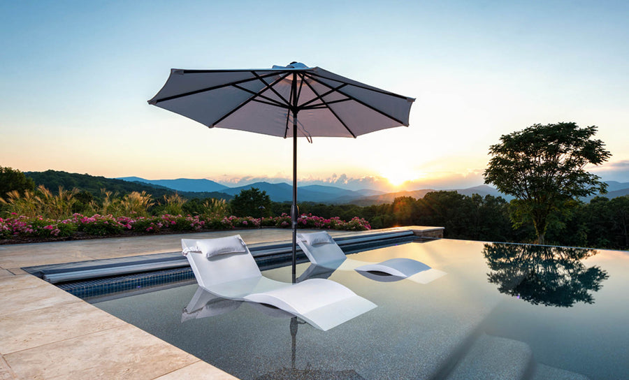 The infinity pool has two submerged loungers with a wind-resistant Ledge Lounger Essential Fiberglass Umbrella providing shade. The scene offers a breathtaking mountain view at sunset, complemented by marine-grade fabrics and vibrant flowers, enhancing the serene ambiance under the clear sky.