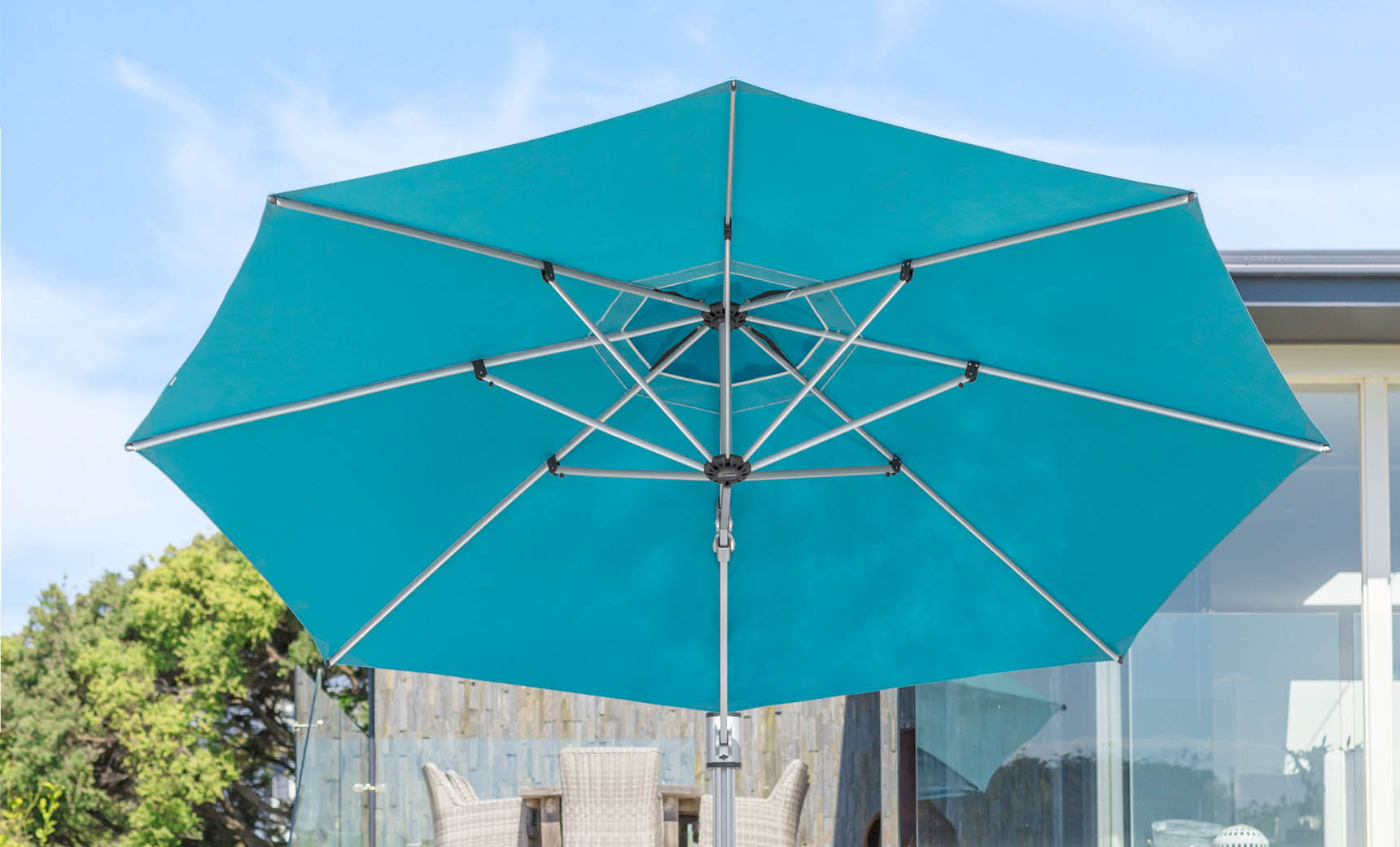 The Ultra Cantilever Umbrella, an 11 octagon by Ledge Lounger, stands open against a clear sky. Its sturdy metal frame casts shade over a welcoming patio with wicker furniture. Trees in the background enhance this serene setting further illuminated by an integrated outdoor light.