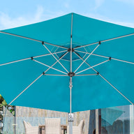The 10 square Ultra Cantilever Umbrella by Ledge Lounger, in turquoise, stands open against a clear sky. Add a rechargeable umbrella light to enhance ambiance and functionality, complementing the hint of patio furniture and lush foliage in the background.