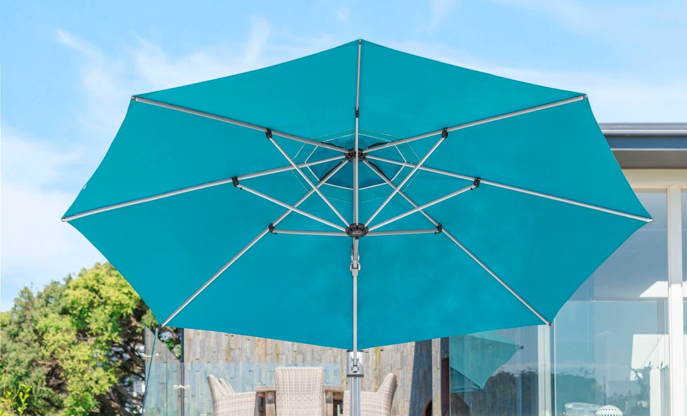 The 10 square Ultra Cantilever Umbrella by Ledge Lounger, in turquoise, stands open against a clear sky. Add a rechargeable umbrella light to enhance ambiance and functionality, complementing the hint of patio furniture and lush foliage in the background.