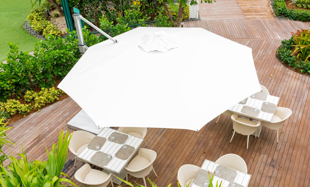 The Ultra Cantilever Umbrella - 10 Square by Ledge Lounger, featuring a rechargeable umbrella light, shelters several square tables and chairs on a wooden deck, surrounded by lush greenery and a manicured lawn for a serene relaxation oasis.