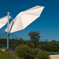 Two Ledge Lounger Ultra Cantilever 10 Square white umbrellas, featuring rechargeable lights, are open by a pool with a clear blue sky backdrop. Glass fencing encloses the pool area, and beyond it, lush green trees and bushes can be seen.