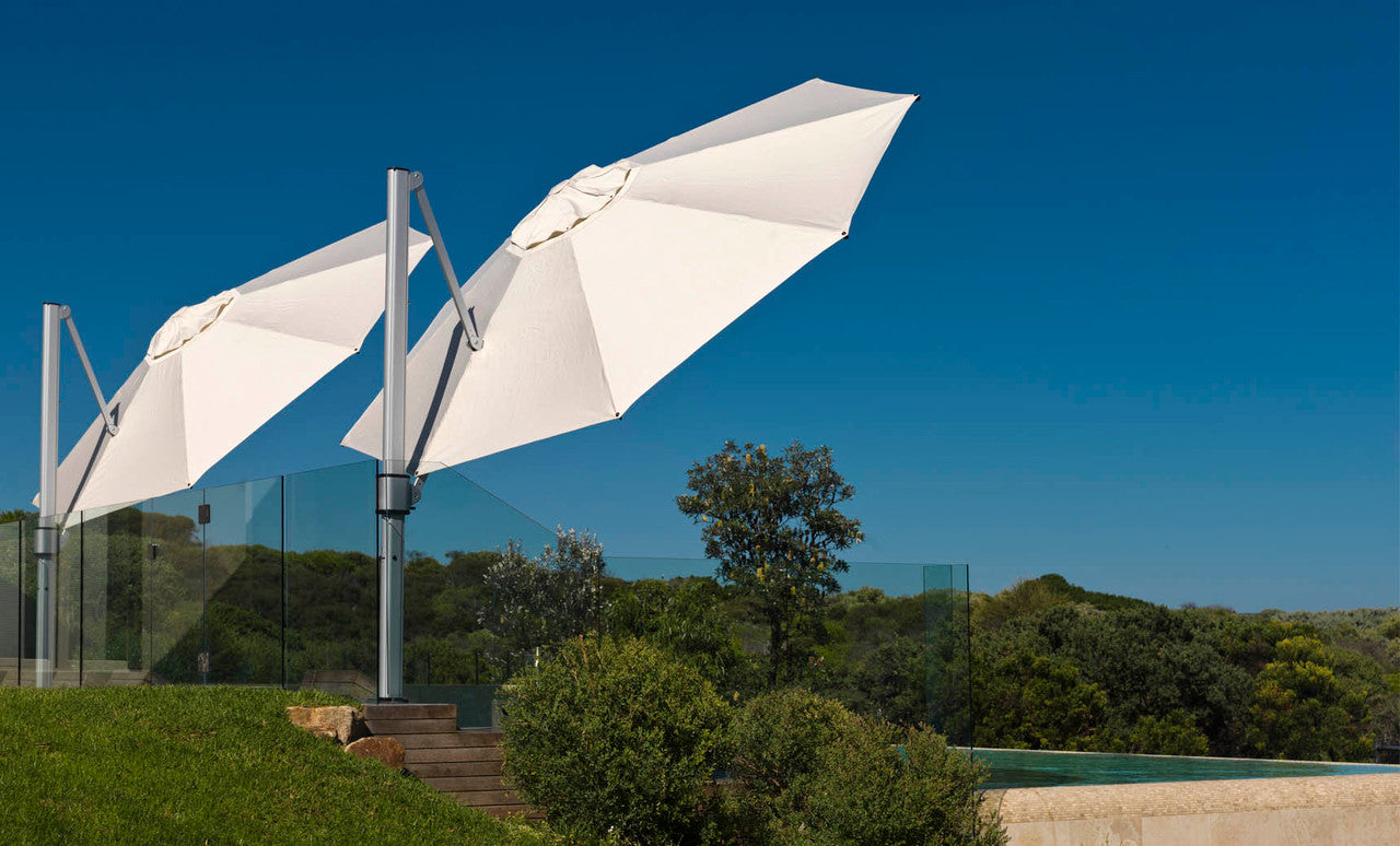 Two Ledge Lounger Ultra Cantilever 10 Square white umbrellas, featuring rechargeable lights, are open by a pool with a clear blue sky backdrop. Glass fencing encloses the pool area, and beyond it, lush green trees and bushes can be seen.